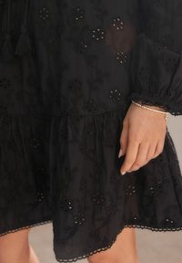 Robe noire brodée avec des motifs floraux, tissu texturé, ourlet volanté et accents de franges. Comprend un bracelet en argent sur une main.