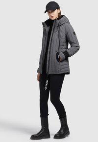 Gray quilted jacket with a hood, black accents, and zipper pockets. Worn with black leggings and sturdy black ankle boots.