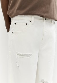 White denim jeans with distressed detailing, featuring frayed rips and black button accents. Straight fit and textured fabric.