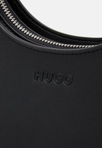 Sort lædertaske med en glat tekstur, subtilt præget logo "HUGO" og en metal lynlåsdetalje.