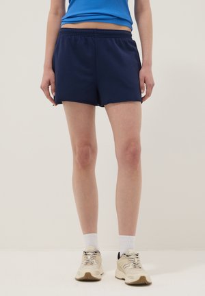 Personne portant un short de sport bleu marine, un haut sans manches bleu, des chaussettes blanches et des baskets beiges, debout devant un fond clair.