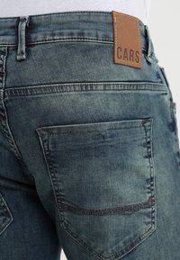 Jeans de mezclilla azul con un aspecto desgastado, que presentan una etiqueta de cuero marrón grabada con "CARS", y dos bolsillos traseros con detalles de costura.
