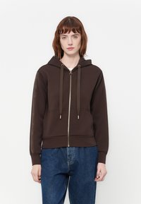 Vrouw met kort rood haar draagt een donkerbruine hoodie met ritssluiting en trekkoorden, samen met een blauwe spijkerbroek, staande tegen een effen witte achtergrond.