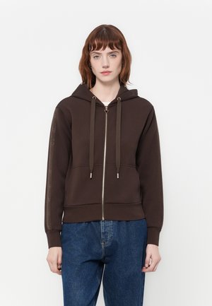 Vrouw met kort rood haar draagt een donkerbruine hoodie met ritssluiting en trekkoorden, samen met een blauwe spijkerbroek, staande tegen een effen witte achtergrond.