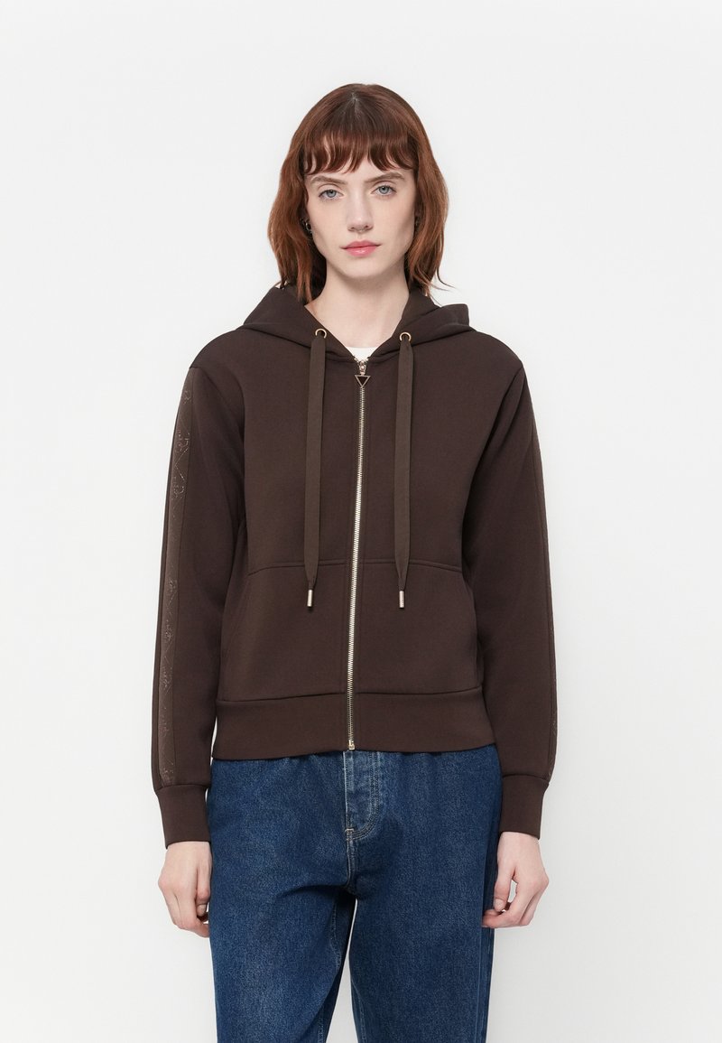 Vrouw met kort rood haar draagt een donkerbruine hoodie met ritssluiting en trekkoorden, samen met een blauwe spijkerbroek, staande tegen een effen witte achtergrond.