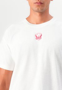 Tricou alb din bumbac, cu guler rotund, având un logo brodat de vultur roșu pe piept. Mâneci scurte, croială lejeră, textură ușoară.