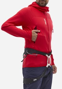 Rote strukturierte Zip-Jacke, die über einem schwarzen Geschirr getragen wird, mit Seitentaschen und einer enganliegenden Kapuze. Die Textur erhöht die Wärme und Beweglichkeit.