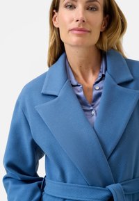 Cappotto in misto lana azzurro chiaro con collo a revers, vita con cintura e texture a coste, indossato sopra una camicia azzurro chiaro con bottoni.