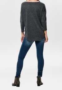 Vrouw in een donkergrijze langslavige top, blauwe skinny jeans en enkellaarzen met een slangenhuidpatroon, met haar rug naar voren gekeerd.