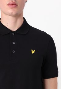 Gros plan d’une personne portant un polo noir avec trois boutons et un petit emblème d’oiseau jaune sur la poitrine gauche.