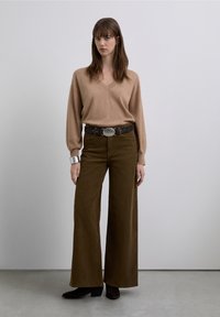 Suéter beige de cuello en V, pantalones anchos de pana marrón, hebilla de cinturón plateada, brazalete ancho grande de plata, botines negros, fondo liso.