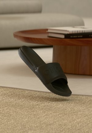 Sandale noire Crocs slide flottant au-dessus d'un tapis beige tissé avec une table basse en bois et un canapé en arrière-plan.