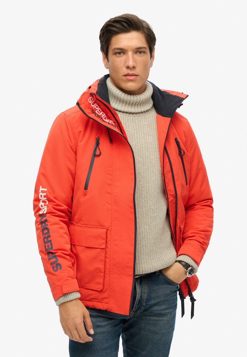 Chaqueta roja acolchada con capucha con cremallera, ribetes negros y detalles de logo. Presenta bolsillos laterales y dobladillo ajustable. Llevada sobre un suéter grueso.