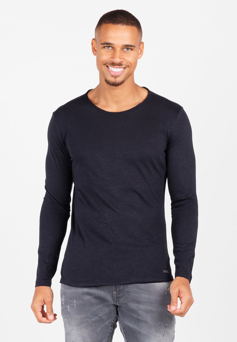 Langzaam, crew-neck t-shirt met lange mouwen in marineblauw, gemaakt van zachte stof. Opvallende stiksels aan de zoom. Gepaard met versleten grijze jeans.