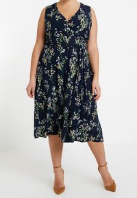 Blommig omlottklänning i marinblått tyg, med midjesnörning, ärmlös design och knälång kjol med en blandning av vita och gula blommor.