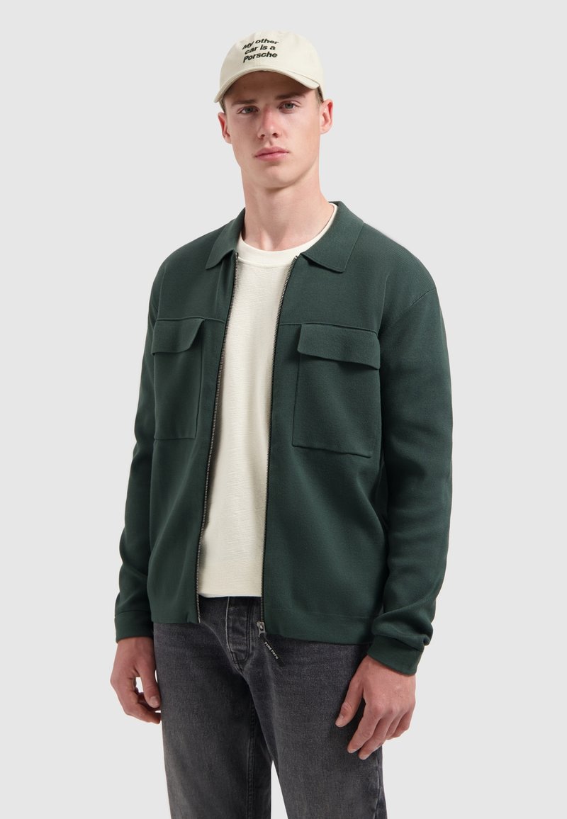 Donkergroene zip-up jack met twee borstzakken, gedragen over een lichtgekleurde shirt, gecombineerd met donkergrijze jeans en een beige pet.