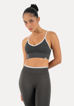 Kobieta z włosami związanymi w koka, ubrana w ciemnoszary sportowy biustonosz z białym wykończeniem i pasujące legginsy z wysokim stanem, stojąca na jednolitym tle.