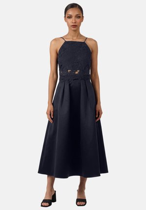 Frau trägt ein ärmelloses marineblaues Spitzenoberteil mit Ausschnitt an der Taille und einen hoch taillierten, plissierten, langen marineblauen Rock mit schwarzen Sandalen mit Absatz.