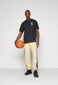 Sort T-shirt med grafisk print, cremefarvede træningsbukser med sorte sidelommer og sorte sneakers. Personen holder en basketball.
