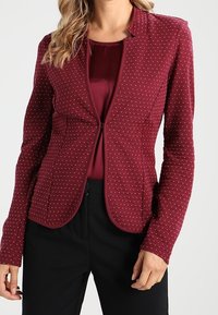 Bordowy żakiet dopasowany z teksturowanym wzorem, z okrągłym dekoltem, subtelnymi przeszyciami i długimi rękawami. Noszony na dopasowanej bluzce.