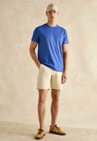 Niebieska bawełniana koszulka, jasnobeżowe szorty, brązowe zamszowe sneakersy z białymi akcentami oraz beżowa czapka z daszkiem. Prosty, casualowy strój.