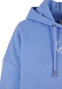 Světle modrá mikina s kapucí z měkké tkaniny. Disponuje stahovací kapucí a vyšitým logem na přední straně. Hladká textura, volný střih.