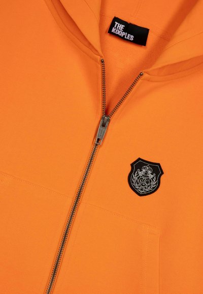 Oranje capuchon sweatshirt met ritssluiting, voorzien van een zilveren rits, voorzakken en een zwarte patch met ingewikkelde zilveren borduursels.