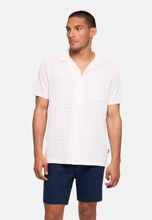 Hombre con camisa blanca texturizada de manga corta con cuello y pantalones cortos azul marino, de pie con expresión neutral mirando hacia la izquierda.