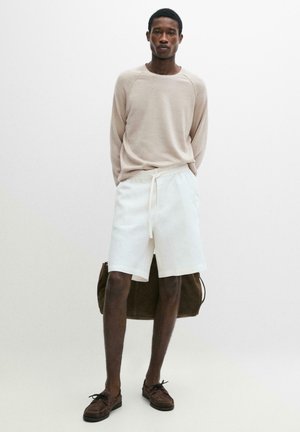 Uomo con camicia beige a maniche lunghe, pantaloncini bianchi con coulisse, mocassini marroni, che tiene una grande borsa marrone dietro di sé su sfondo bianco.