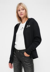 Čierna mikina na zips s logom na hrudi, nosená cez biely tričko. Tmavé, textúrované slim-fit nohavice dotvárajú outfit.