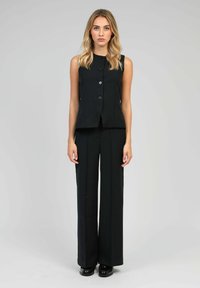 Femme aux cheveux blonds portant un gilet sans manches noir à boutons, un pantalon large noir et des mocassins noirs, se tenant devant un fond gris clair.