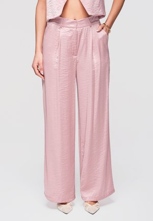 Kobieta nosząca błyszczące różowe spodnie z wysokim stanem i szerokimi nogawkami, z kieszeniami po bokach, oraz białe plecione szpilki z ostro zakończonymi noskami, stojąca na jednolitym tle.