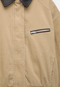Jachetă din înveliș de corduroy bej cu guler din piele ecologică neagră, având un buzunar cu fermoar pe piept și o textură verticală cu nervuri.