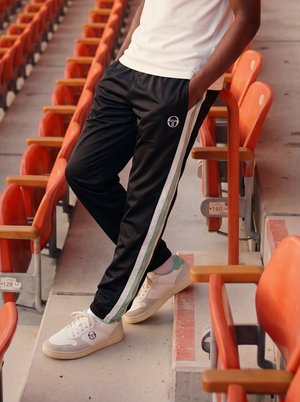Person iført sorte træningsbukser med hvide striber og hvide sneakers står på tomme orange stadionpladser.