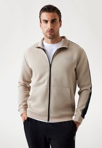 Giacca beige con zip e colletto rialzato, con accenti neri laterali e un logo sul davanti. Tessuto morbido e testurizzato, abbinato a una maglietta bianca sotto.