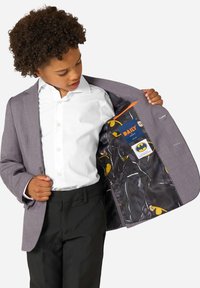 Chłopiec w eleganckim stroju rozkłada szary blezer, aby pokazać podszewkę z motywem Batmana, na której znajdują się żółte loga i postacie w kieszeni.