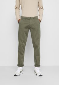 Olijfgroene chinos met een tailored fit, voorzien van zijzakken en opgerolde manchetten, gecombineerd met witte en grijze sportschoenen.
