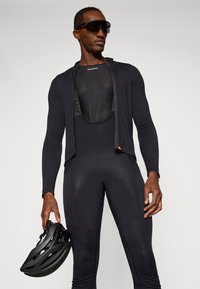 Îmbrăcăminte de ciclism neagră, care include un tricou cu mâneci lungi și fermoar în față, alături de colanți cu padding. O cască neagră este ținută în mâna stângă.