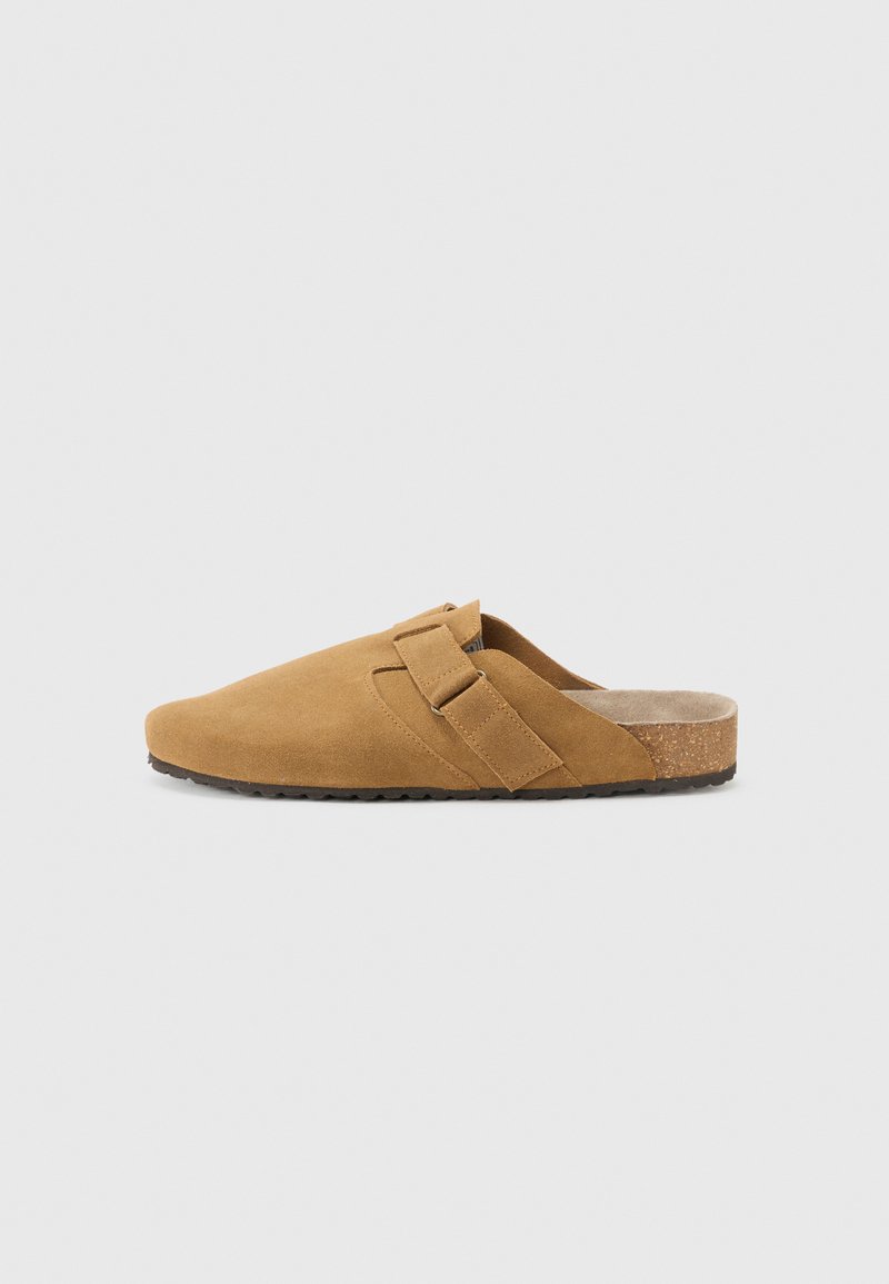 Brązowe zamszowe buty typu slip-on z szerokim paskiem na górze, teksturowaną wkładką i korkową podeszwą środkową. Płaska gumowa podeszwa zewnętrzna zapewnia przyczepność.