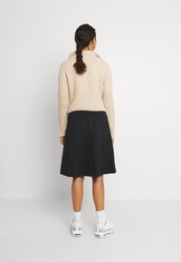 Béžový pletený svetr s vysokým límcem, doplněný černou, strukturovanou sukní po kolena. Bílé tenisky a kotníkové ponožky dotvářejí celkový vzhled.