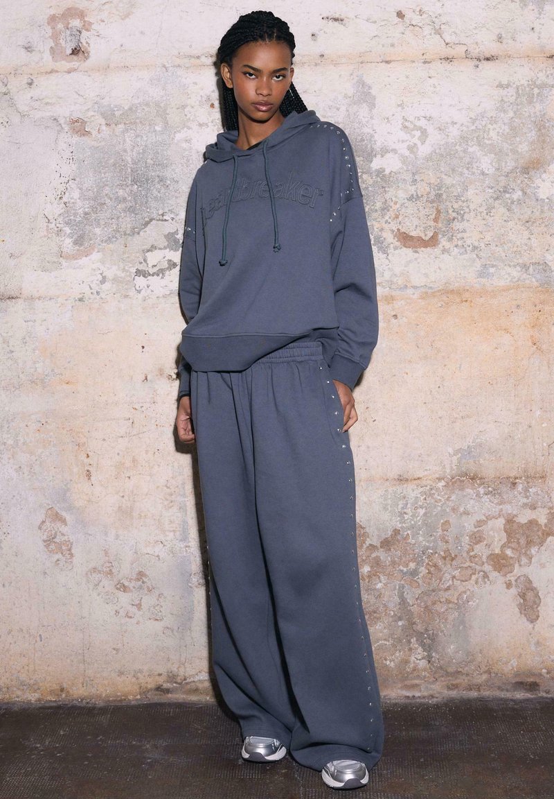 Jeune femme aux cheveux tressés portant un sweat à capuche ample gris foncé et un pantalon large orné de détails cloutés, se tenant contre un mur usé et texturé.