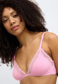 Růžová trojúhelníková podprsenka s krajkovou ozdobou po okrajích, nastavitelné ramínka a hladká textura látky. Není vidět žádná kostice.