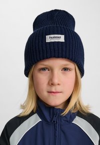 Mörkblå stickad mössa med uppvikt brätte, prydd med en vit logotyp. Bärs tillsammans med en blå och grå sportjacka.