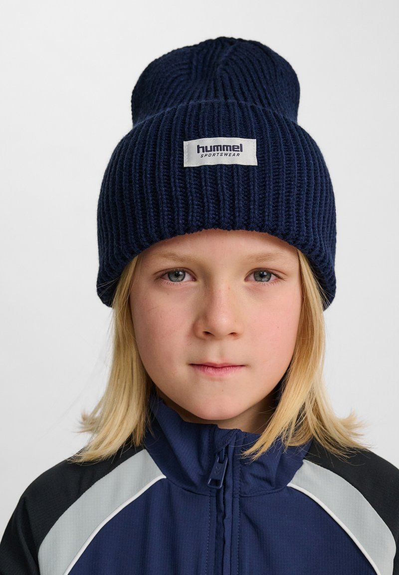 Mörkblå stickad mössa med uppvikt brätte, prydd med en vit logotyp. Bärs tillsammans med en blå och grå sportjacka.