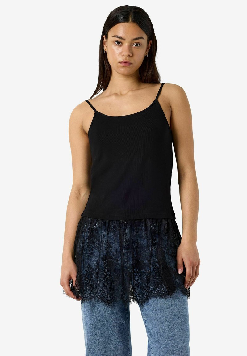 Jeune femme portant un haut noir à fines bretelles avec un overlay en dentelle noire attaché et un jean bleu, se tenant devant un fond blanc.