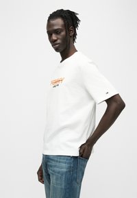 Λευκό βαμβακερό T-shirt με πορτοκαλί τυπωμένο κείμενο "TOMMY JEANS." Διαθέτει κοντά μανίκια και κλασικό στρογγυλό λαιμό. Φοριέται με μπλε τζιν παντελόνια.