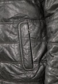 Nahaufnahme einer schwarzen Lederjackentasche mit sichtbaren Nähten und teilweise freiliegendem Metallreißverschluss.