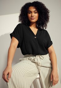 Top nero a maniche corte con scollo a V e dettagli con bottoni, abbinato a pantaloni crema a righe con vita regolabile. Texture morbida.