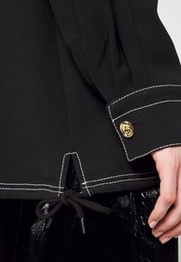 Manche d'un vêtement noir avec des coutures blanches et un bouton doré au poignet, présentant un cordon noué à l'ourlet et une main lisse reposant à côté.