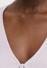 Tejido de color púrpura claro con diseño de escote en V y un pequeño detalle de nudo en la parte delantera. Textura suave contra el tono de piel.